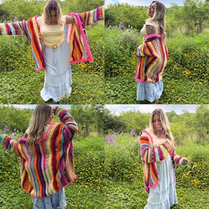 May include: A colorful, handmade crochet cardigan with a striped pattern in shades of pink, orange, yellow, green, and purple. The cardigan has long sleeves and a bright pink trim. The model is wearing a light blue dress.