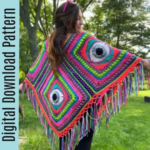 Op de afbeelding: Een kleurrijk gehaakt poncho met een franje rand en oogontwerpen. De poncho is gemaakt met fel roze, groen, blauw en paars garen.