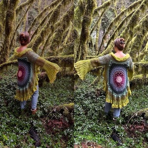 May include: A person wearing a crocheted cardigan with a colorful mandala design in the back. The cardigan has long sleeves and a flowing hem. The person is standing in a forest setting with moss-covered trees.