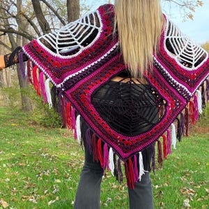 May include: A handmade crochet poncho in shades of white, purple, pink, and red, featuring a spiderweb design on the shoulders and a fringed hem. The poncho is worn over a black top and dark gray pants, with a natural outdoor background.