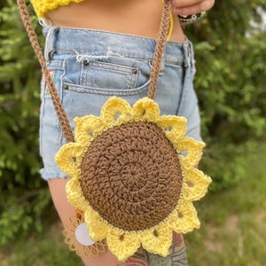 May include: A handmade crochet sunflower bag with a brown centre and yellow petals. The bag has a brown strap and a small sunflower charm. The bag is worn over the shoulder.