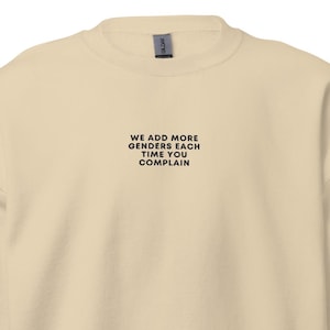 May include: A cream-colored sweatshirt with the text "WE ADD MORE GENDERS EACH TIME YOU COMPLAIN" in black. The sweatshirt has a crew neck and long sleeves with ribbed cuffs and hem.