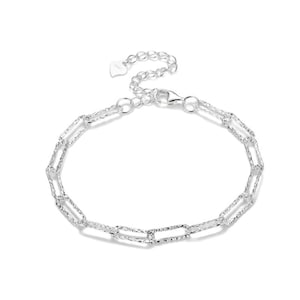Könnte beinhalten: Ein silbernes Kettenarmband mit strukturiertem Design. Das Armband hat einen Karabinerverschluss und einen herzförmigen Anhänger.