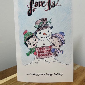 May include: A hand-drawn Christmas card featuring a snowman with two friends. The snowman is wearing a brown hat with a green sprig. The friends are wearing colorful hats and scarves. The snowman is holding a sign that says "Happy Holidays From Us" with a heart on each side of the sign. The card says "Love is... wishing you a happy holiday."