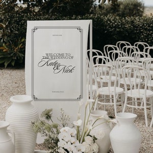 Puede incluir: Un cartel de boda blanco con texto negro que dice "Bienvenido a la boda de Kate & Nick" con la fecha y la ubicación debajo. El cartel está de pie frente a una fila de sillas blancas y un arreglo floral con flores blancas.