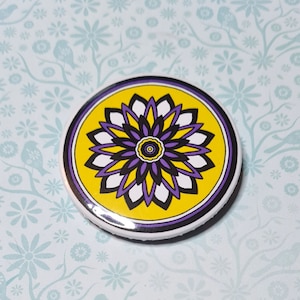 Può includere: Un bottone rotondo con un design a mandala giallo, viola e nero. Il mandala ha un cerchio centrale con un design giallo e viola, circondato da un design bianco e nero.