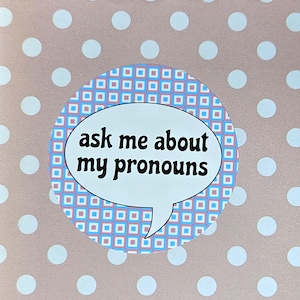 Pode incluir: Um fundo rosa e branco de bolinhas com um círculo estampado em azul e rosa. O círculo tem um balão de fala branco com o texto "ask me about my pronouns".