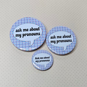 May include: Three round buttons with a pink and blue checkered pattern. The buttons have a white speech bubble with the text "ask me about my pronouns" in black.