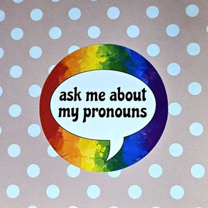 Peut inclure: Un cercle arc-en-ciel avec une bulle de dialogue blanche qui dit "ask me about my pronouns".