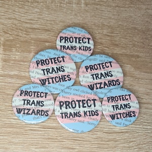 Op de afbeelding: Vijf ronde knoppen met een roze, wit en blauw kleurenschema. Elke knop heeft de tekst "Protect Trans" gevolgd door een ander woord: "Kids", "Witches", "Wizards", "Kids" en "Witches".
