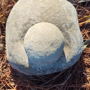 Pode incluir: Um almofariz de pedra cinza claro com um pilão esférico menor. O almofariz tem uma forma grande e irregular, e o pilão está em cima. Os objetos estão sobre uma cama de agulhas de pinheiro marrons.