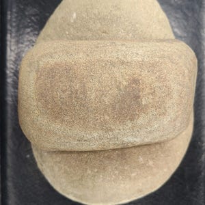 Pode incluir: Um artefato de pedra castanho claro e desgastado com uma forma arredondada única. A pedra tem uma superfície texturizada e uma faixa no meio. A forma geral sugere uma ferramenta antiga ou uma escultura.