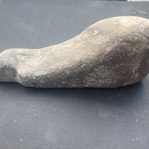 May include: A smooth, grey stone with a rounded head and a curved body. The stone has a natural, weathered appearance, with variations in colour and texture. The object is likely a decorative piece or a natural artefact.