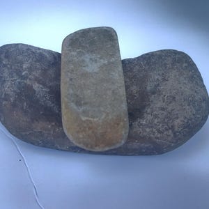 Pode incluir: Três pedras naturais dispostas juntas. Uma pedra retangular castanho-clara está centrada entre duas pedras arredondadas maiores, cinza escuro. As pedras têm uma superfície áspera e texturizada.