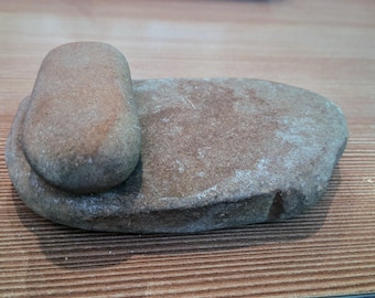 Puede incluir: Un juego de mortero y mano de piedra natural que presenta una base de molienda plana de forma ovalada y una herramienta de mano redondeada y lisa. Las piedras son de color marrón tierra y gris con una superficie texturizada y desgastada.