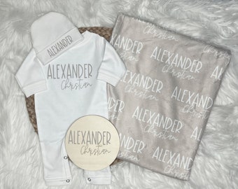 May include: A white baby onesie with the name "Alexander Christian" printed on it. A white beanie with the same name is included. A beige blanket with the name "Alexander Christian" printed on it is also included. A wooden round sign with the same name is included.