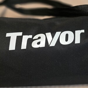 May include: Black bag with the white word "Travor" printed on the front. The bag has a black strap and appears to be made of a durable material. The text is in a bold, sans-serif font.