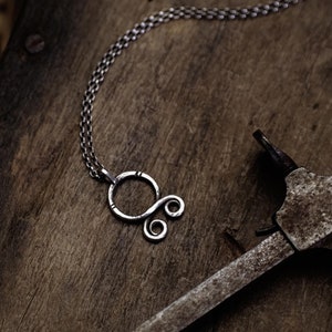 May include: A silver necklace with a unique, abstract pendant. The pendant is made of a single piece of wire that is twisted and shaped into a circle with a smaller circle and two loops coming off of it. The necklace is lying on a rustic wooden surface.
