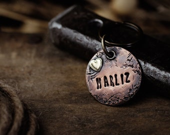 May include: A copper dog tag with the name "Marliz" engraved on it. A small gold heart charm is attached to the tag.