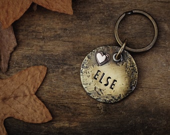 May include: A brass dog tag with the name "ELSE" engraved on it. A small heart charm is attached to the tag. The tag is on a wooden surface with fall leaves.