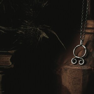 May include: A silver pendant necklace with a circular design featuring a knot or symbol. The necklace is hanging from a brown leather surface.