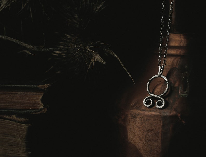 May include: A silver pendant necklace with a circular design featuring a knot or symbol. The necklace is hanging from a brown leather surface.