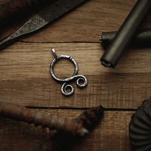 May include: A silver metal pendant with a circular design and two smaller circles. The pendant is on a wooden surface with other metal objects.