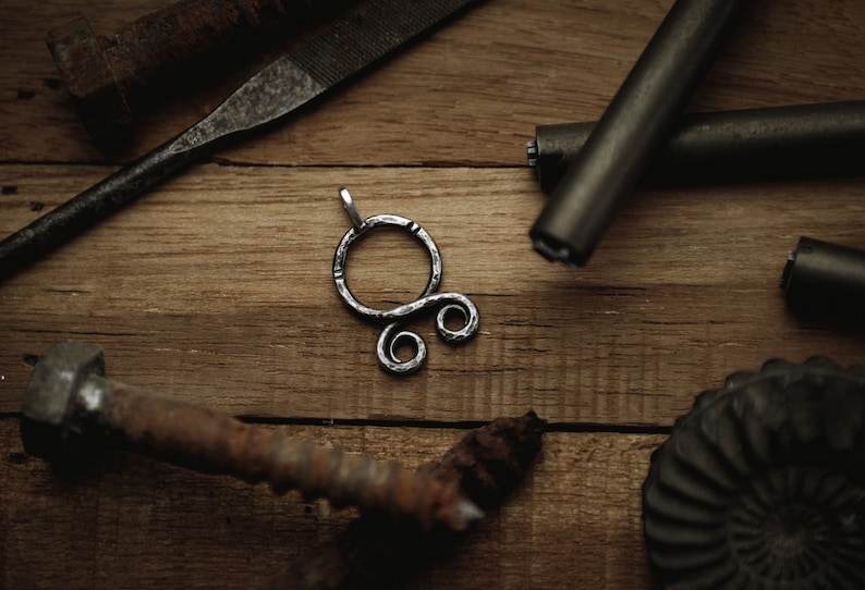 May include: A silver metal pendant with a circular design and two smaller circles. The pendant is on a wooden surface with other metal objects.