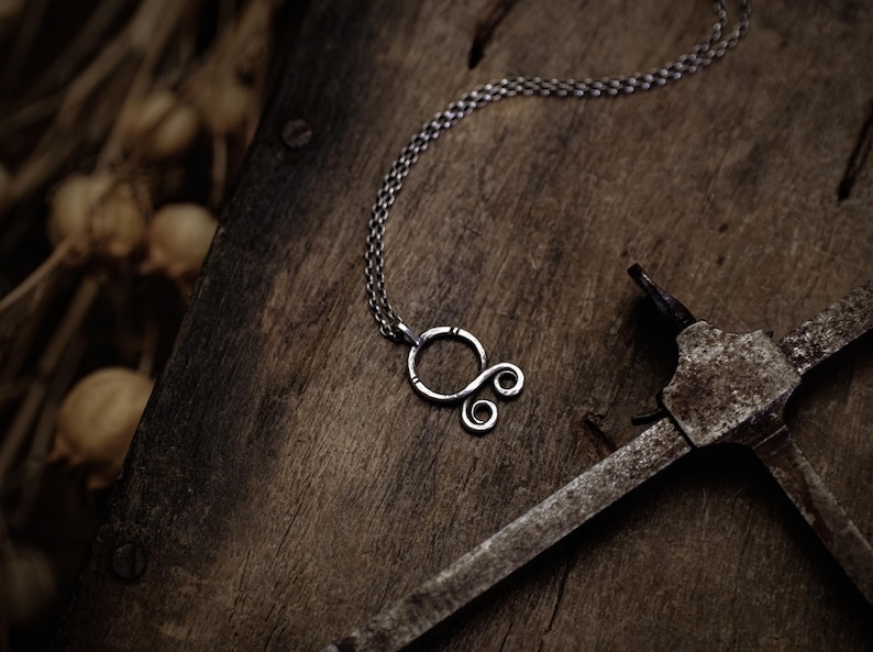 May include: A silver necklace with a unique, abstract pendant. The pendant is made of a single piece of wire that is twisted and shaped into a circle with a smaller circle and two loops coming off of it. The necklace is lying on a rustic wooden surface.