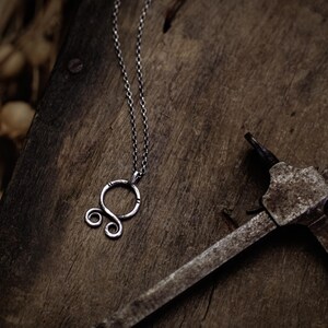 May include: A silver necklace with a circular pendant featuring a stylized double spiral design. The necklace is displayed on a rustic wooden surface.