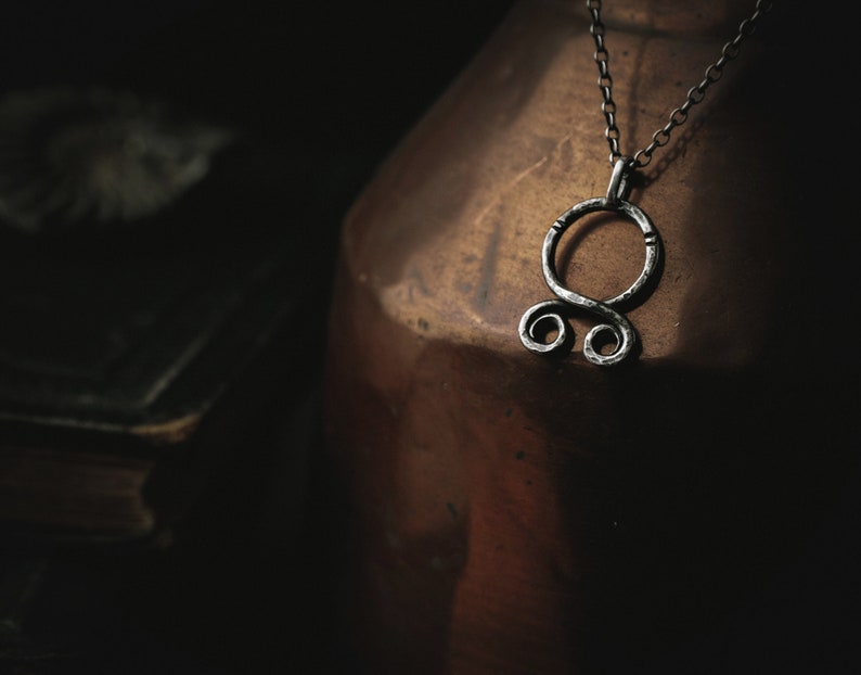 May include: A silver pendant necklace with a stylized knot design hangs from a silver chain. The necklace is resting on a brown ceramic vase.