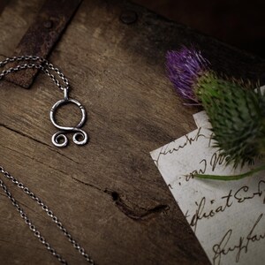 May include: A silver necklace with a Celtic knot pendant on a wooden surface. The pendant is a circle with two smaller circles on the bottom. The necklace is on a wooden surface with a piece of paper with handwritten text.