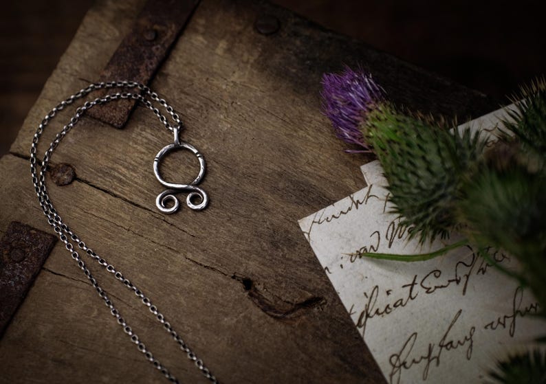 May include: A silver necklace with a Celtic knot pendant on a wooden surface. The pendant is a circle with two smaller circles on the bottom. The necklace is on a wooden surface with a piece of paper with handwritten text.