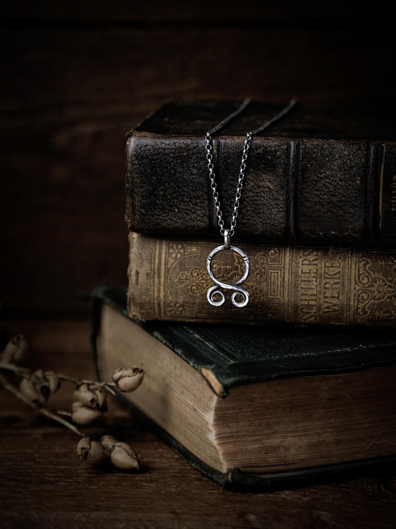May include: A silver necklace with a circular pendant featuring a stylized double-loop design. The necklace is resting on top of a stack of antique books.