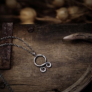 May include: A silver chain necklace with a silver pendant featuring a stylized knot design. The necklace is lying on a wooden surface with a piece of antler in the background.