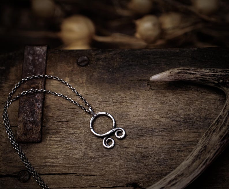 May include: A silver chain necklace with a silver pendant featuring a stylized knot design. The necklace is lying on a wooden surface with a piece of antler in the background.