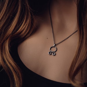 May include: A silver necklace with a unique pendant featuring a circle with two smaller circles attached to the bottom. The pendant is made of textured metal.
