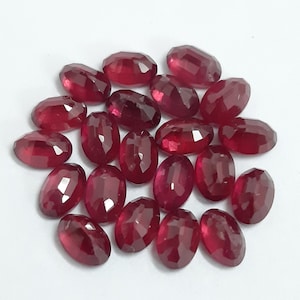 May include: A group of 20 oval-shaped, faceted, red gemstones. The gemstones are arranged in a loose pile on a white background.