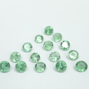 Puede incluir: Una colección de 15 piedras preciosas redondas, facetadas y verdes. Las piedras preciosas están dispuestas en un patrón triangular suelto sobre un fondo blanco.