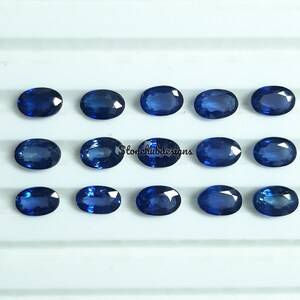Könnte beinhalten: Eine Sammlung von 15 ovalen blauen Edelsteinen, wahrscheinlich Saphiren, die in drei Reihen auf einer weißen Oberfläche angeordnet sind. Die Edelsteine sind alle ungefähr gleich groß und geformt.