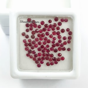 May include: A collection of small, round, deep red gemstones, possibly rubies, are displayed inside a white square container. The gemstones are scattered across a white surface. The text "Stonehubdesigns" is visible on the container.