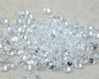 May include: A collection of small, clear, faceted gemstones scattered on a light beige surface. The gemstones are round and reflect light, creating a sparkling effect. The image is a close-up, showcasing the details of the stones.