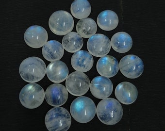 Puede incluir: Colección de cabujones redondos de piedra lunar pulida. Las piedras son translúcidas con una base blanca lechosa y exhiben adularescencia azul. Los cabujones están dispuestos sobre una superficie negra. El texto "Stonehubdesigns" es visible.