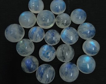 Puede incluir: Una colección de 17 cabujones de piedra lunar blanca con destellos azules iridiscentes. Los cabujones son redondos y lisos, y están dispuestos en un patrón aleatorio.