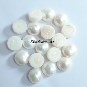 May include: A selection of white, half-drilled, round pearls with a glossy sheen. Some pearls have a flat back, while others have a drilled hole. The pearls are scattered on a white surface. The text "Stonehubdesigns" is visible.