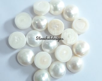 May include: A collection of white, half-drilled, round pearls with a lustrous sheen. Some pearls have a flat back, while others have a drilled hole. The pearls are scattered on a white surface. The text "Stonehubdesigns" is visible.