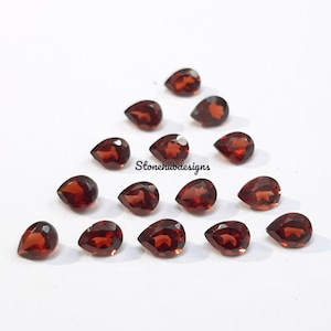 Puede incluir: Una colección de 15 piedras preciosas rojas en forma de pera, probablemente granates, dispuestas en un patrón triangular sobre una superficie blanca. Las piedras preciosas son todas de tamaño y forma aproximadamente iguales.