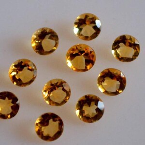 May include: Ten round, faceted, golden yellow gemstones. The gemstones are arranged in a scattered pattern on a white surface.