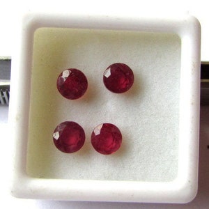 Puede incluir: Cuatro piedras preciosas redondas de color rojo en una caja cuadrada blanca. Las piedras preciosas son facetadas y tienen un acabado brillante.