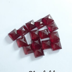 May include: A group of 14 red square-shaped glass gems. The gems are faceted and have a shiny finish.